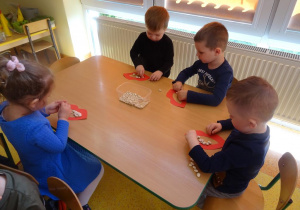 Dzieci siedzą przy stoliku, układają uśmiech z wykorzystaniem szablonu ust i ziaren fasoli.